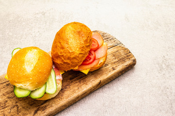 Homemade sandwich in bun. Salami, dried pork, tomato, cucumber, cheese ingredients. On wooden board and stone background.