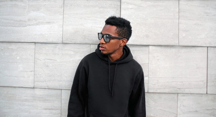 Portrait african man in black hoodie, sunglasses looking away on city street over gray brick wall background © guas