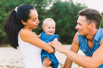 A young, beautiful family of three. Mom, Dad and daughter in the arms of my father play, rejoice, smile on the sandy beach on the beach in the summer. Dressed in junky clothes and white shirts