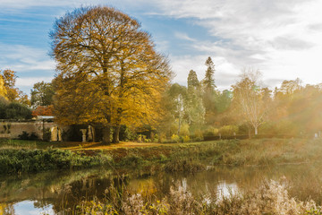Beautiful autumn garden with tree reflections in water, blue sky, sun flares. Hever Castle Gardens,...