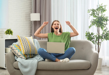Emotional young woman with laptop celebrating victory on sofa at home