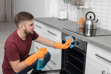 Young man cleaning oven with rag and detergent in kitchen