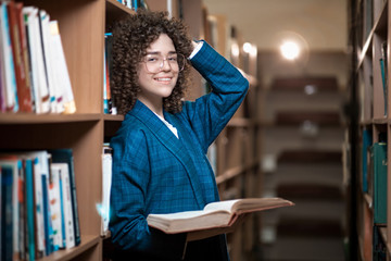 Young beautiful curly girl in glasses and a blue suit is standing in the library. Student Study