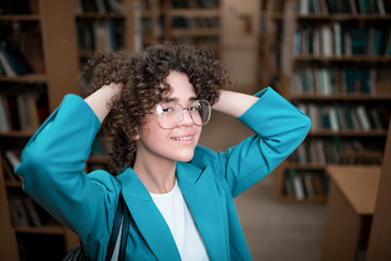 Young beautiful curly girl in glasses and a blue suit is standing in the library. Student Study