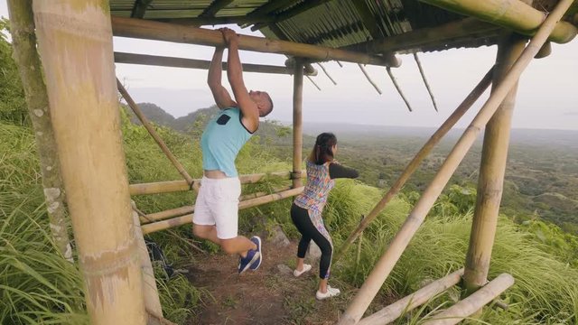 Fitness couple training sport exercise together on outdoor ground on natural landscape. Man doing pull up exercise outdoor. Fit woman training squat exercise on tropical landscape.
