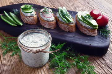 glass jar with feta, cottage cheese, and dill spread and sandwiches with raw vegetables and the spread on black serving board on wooden table decorated with dill