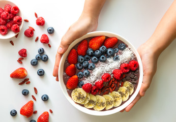 Woman hands holding an Acai Berry superfoods smoothies white bowl with chia seeds, strawberry, goji berry, coconut, blueberry, raspberry toppings, and chocolate chip on white background