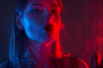 Glamorous brown-haired woman breathes on a window with water drops in neon light close-up. - 258250603