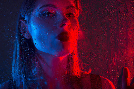Glamorous brown-haired woman breathes on a window with water drops in neon light close-up.
