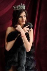 close up photo of a sexy fashion woman in black fur coat with a crown on her head on a red curtain background in a photo studio