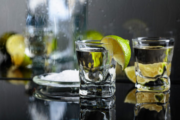 Glasses of tequila with lime and salt on the black reflective background.