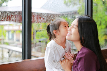 Adorable little Asian child girl kissing her mother with love near a glass window with water drop while raining day.