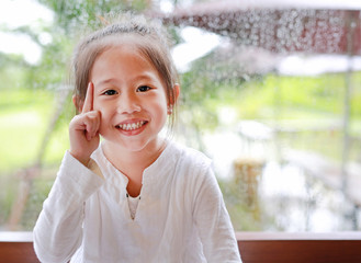 Adorable little Asian child girl gesture with positive funny face against glass window with water drop at rainy day.