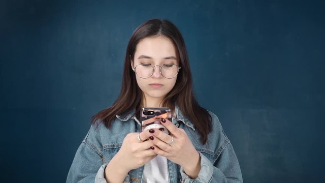 Attractive young girl with glasses typing a message on the phone, smiling, looking at the phone screen. Brunette in denim jacket on blue wall background. Close-up portrait. Slow motion. HD