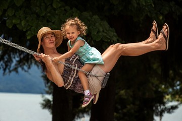Happy loving family! Young mother and her child daughter swinging on the swings and laughing a summer evening outdoors, beautiful girl on the swing in the park by lake, joyful holidays