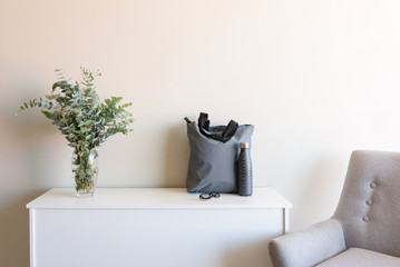 White sideboard with eucalyptus leavesin glass vase, grey shopping tote and reusable drink bottle...