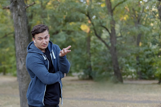Young handsome caucasian man with perplexed face expression and skeptical gesture at green park, wearing casual. Outdoors, copy space, strong emotions.