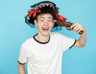 young funny male teenager in white t-shirt on blue background in pirate hat and gun in hand wants to shoot in head