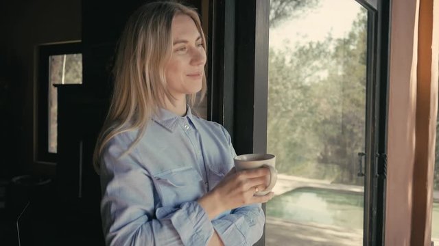 Female drinking coffee in sunny morning at home near big window. Blonde woman with cup of hot tea.