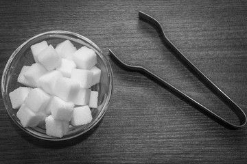 Top view of sugar cubes in a glass bowl with a pair of kitchen tongs foreceps kept on wooden floor. Contrasty black and white kitchen setup.