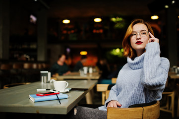 Cheerful young beautiful redhaired woman in glasses sitting at her working place on cafe with cup of coffee.