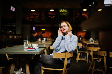 Cheerful young beautiful redhaired woman in glasses sitting at her working place on cafe with cup of coffee.