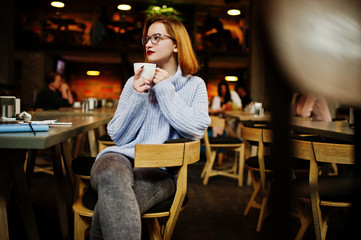 Cheerful young beautiful redhaired woman in glasses sitting at her working place on cafe and drinking coffee.