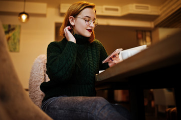 Cheerful young beautiful redhaired woman in glasses, green warm wool sweater, using her phone and notebook, while sitting at her working place on cafe.