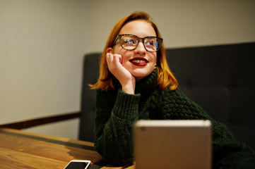 Cheerful young beautiful redhaired woman in glasses, wear on green warm wool sweater,  using her...