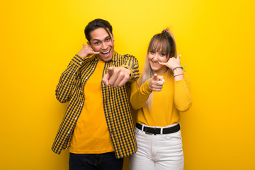 Young couple over vibrant yellow background making phone gesture and pointing front