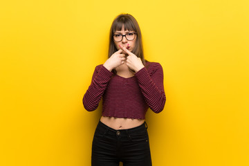 Woman with glasses over yellow wall showing a sign of silence gesture