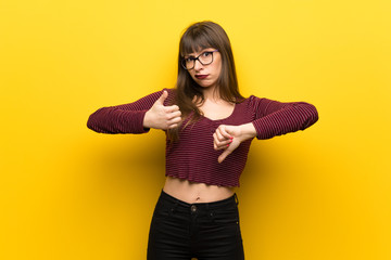 Woman with glasses over yellow wall making good-bad sign. Undecided between yes or not