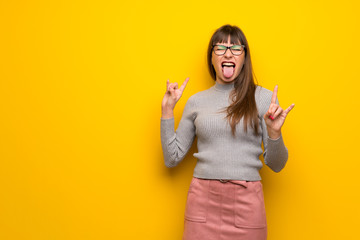Woman with glasses over yellow wall making rock gesture