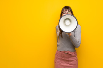Woman with glasses over yellow wall shouting through a megaphone to announce something