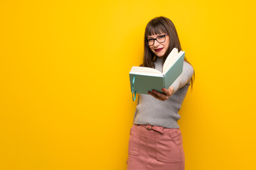 Woman with glasses over yellow wall holding a book and giving it to someone