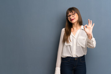 Woman with glasses over blue wall showing an ok sign with fingers