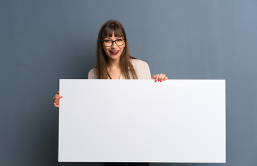 Woman with glasses over blue wall holding an empty placard for insert a concept