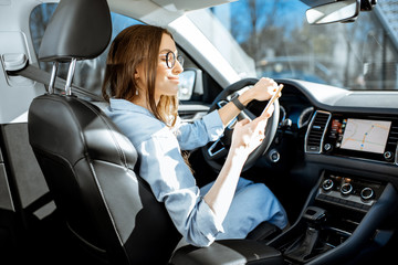 Young and cheerful woman using smart phone while driving a modern car in the city