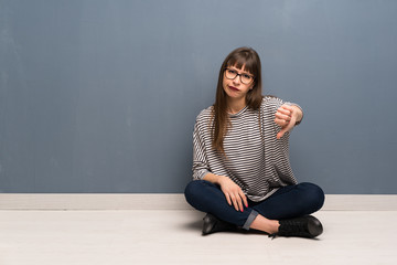 Woman with glasses sitting on the floor showing thumb down with negative expression