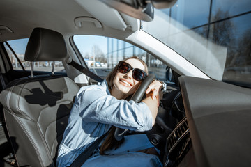 Young and cheerful woman enjoying new car hugging steering wheel sitting inside, wide angle view