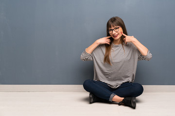 Woman with glasses sitting on the floor smiling with a happy and pleasant expression