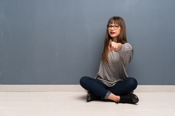 Woman with glasses sitting on the floor points finger at you with a confident expression