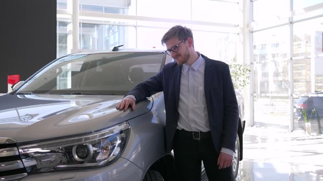 auto purchase, young buyer man with glasses gladly strokes new car and gives positive gesture in showroom