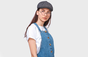 Studio image of pretty young woman wears transparent trendy eyewear, casual white t-shirt, denim dress, trendy gray cap, looks seriously directly into camera, poses on white wall. People emotions