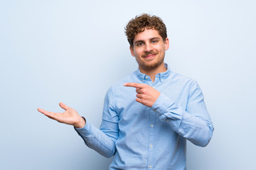 Blonde man over blue wall holding copyspace imaginary on the palm to insert an ad