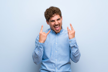 Blonde man over blue wall making rock gesture