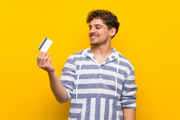 Blonde man over yellow wall holding a credit card and thinking