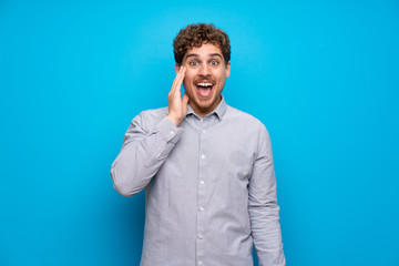 Blonde man over blue wall with surprise and shocked facial expression