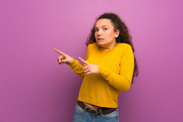 Teenager girl over purple wall frightened and pointing to the side