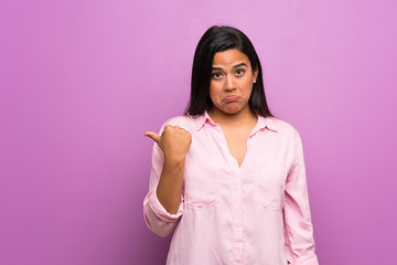 Young Colombian girl over purple wall unhappy and pointing to the side